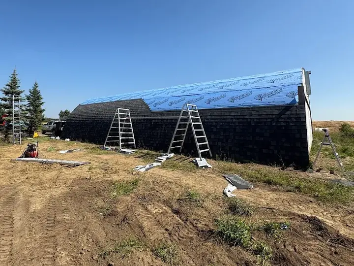 A quonset mid-job, shingled along the lower sides with blue underlayment over the top and ladders against the wall